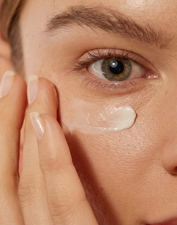 A close-up of a person's fingers applying a white cream under their eye.