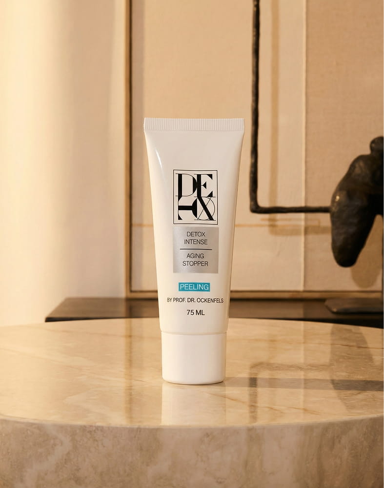 A white tube of Detox Intense Peeling cream stands upright on a marble table against a neutral background.