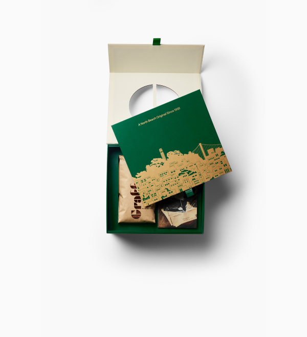 An open green gift box containing a bag of coffee and a card with a city skyline.