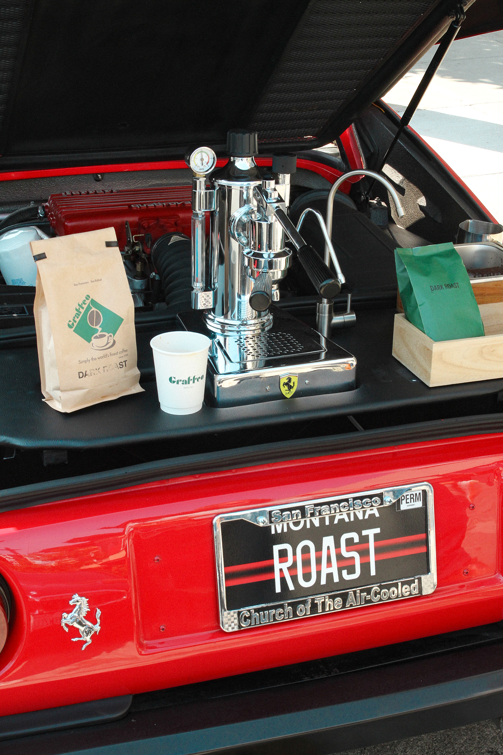 Espresso machine setup in the trunk of a red sports car with 'ROAST' license plate.