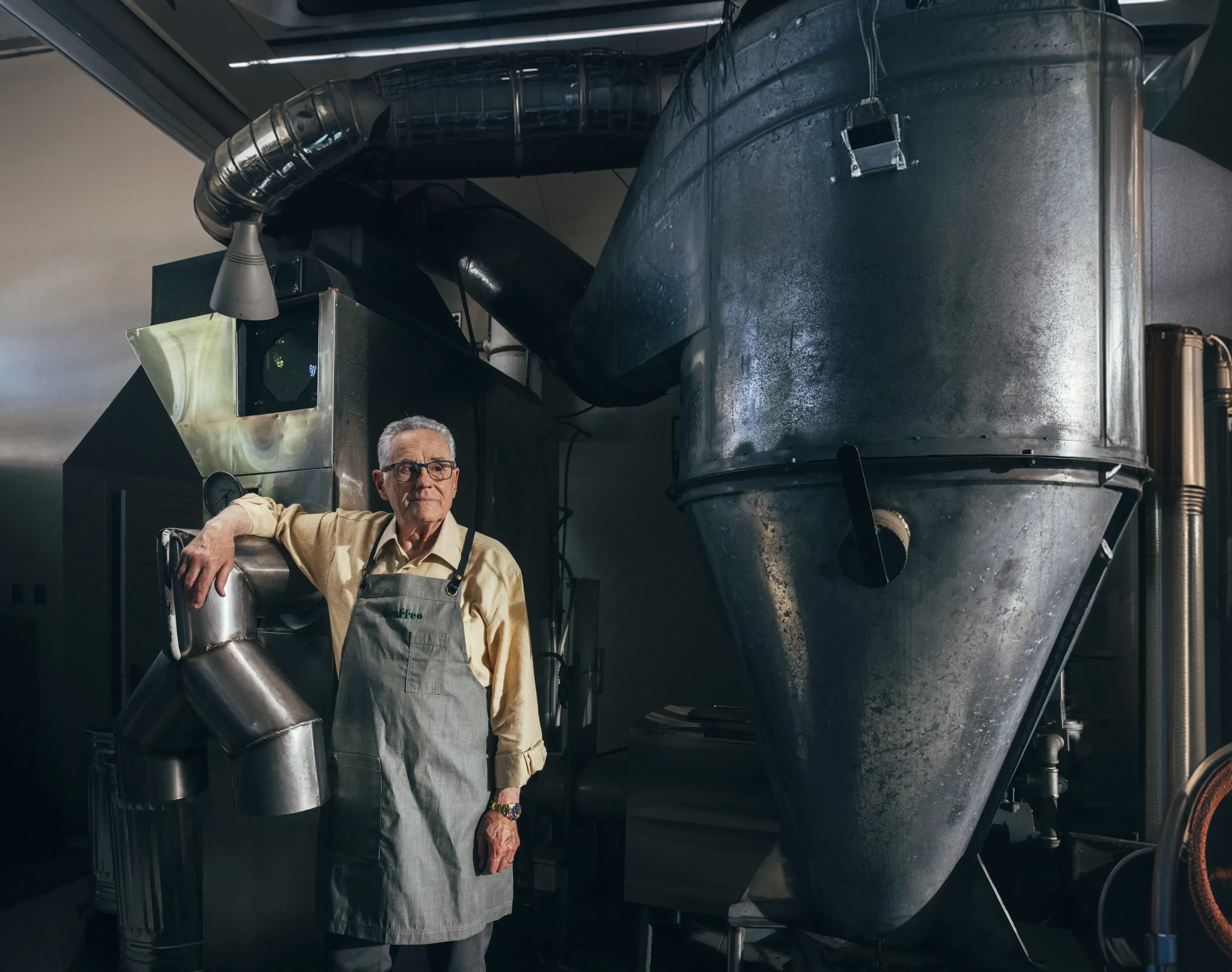An older man in an apron stands next to a large piece of industrial metal machinery.