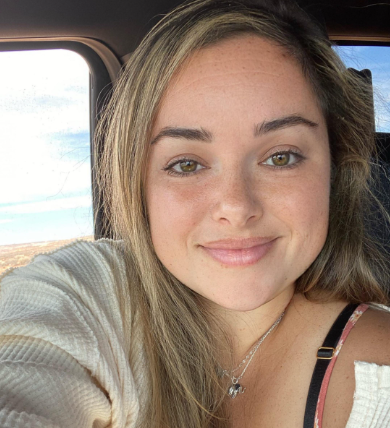 A close-up selfie of a young woman with long, light brown hair and hazel eyes smiling.