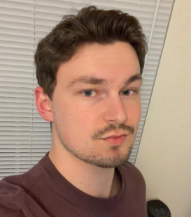 A headshot of a young man with brown hair and a mustache, looking at the camera.