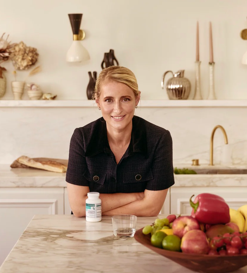 Woman smiling in kitchen with fruits and a supplement bottle.