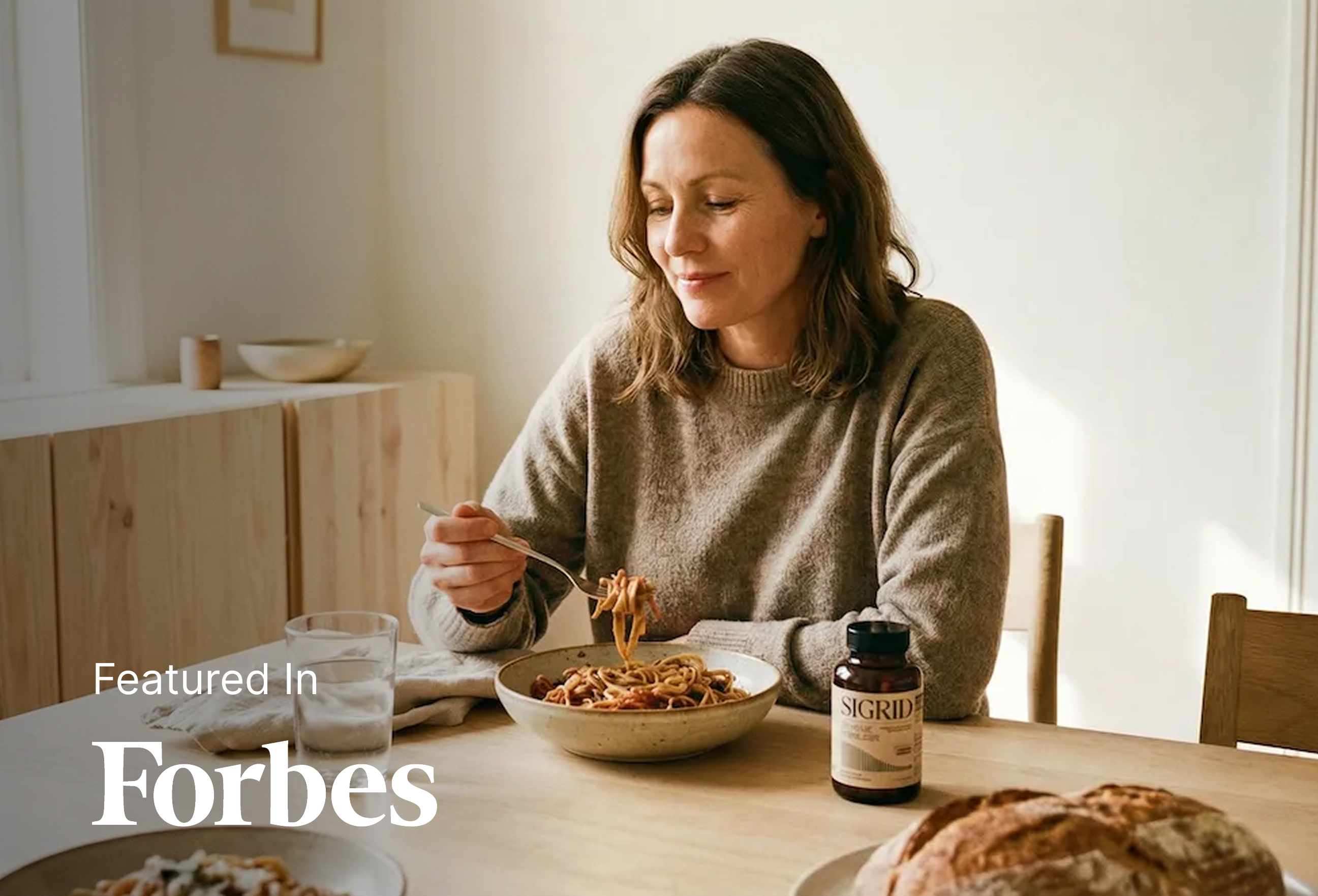 A woman in a brown sweater eats a bowl of pasta at a wooden table with a supplement bottle.