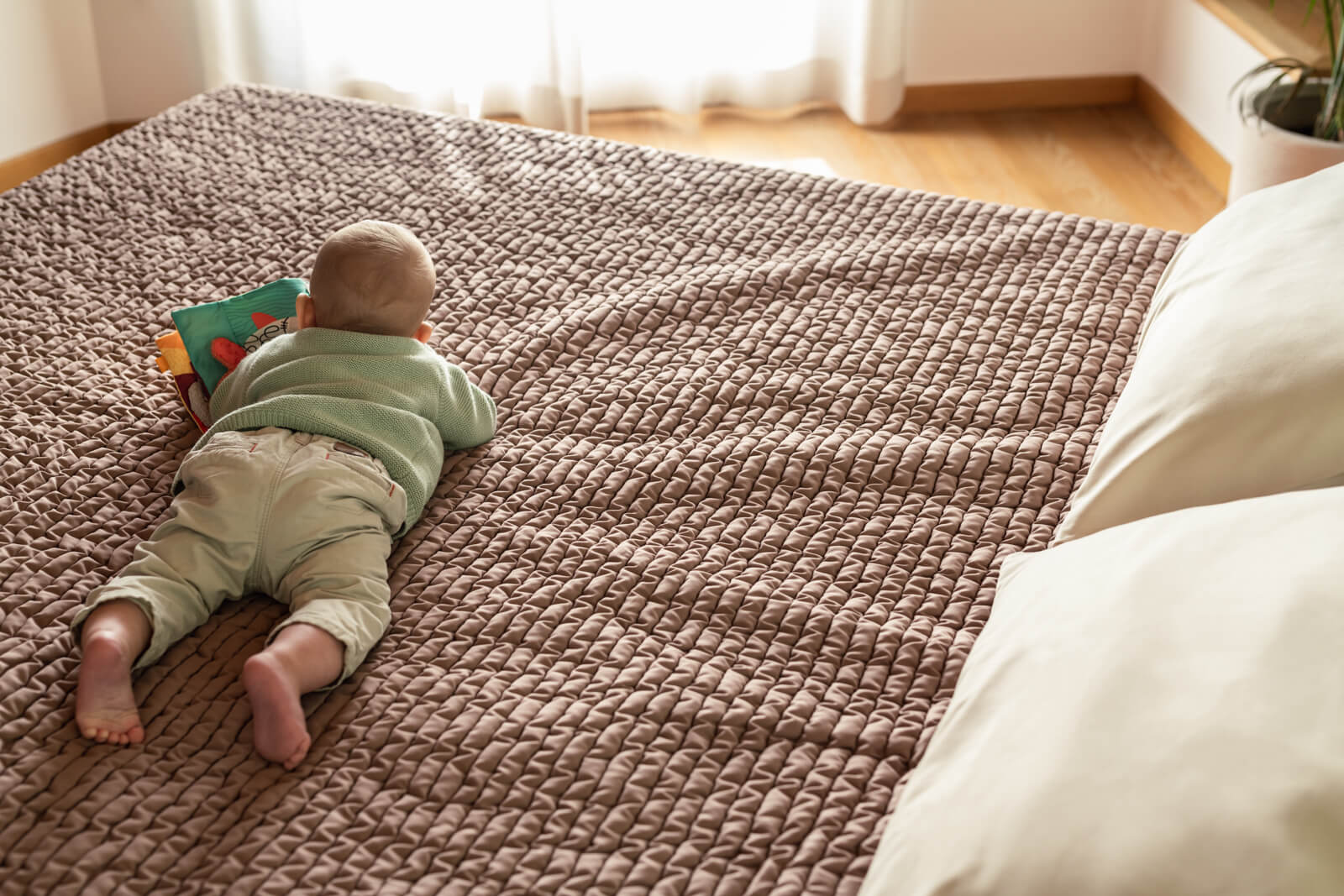 A baby lies on its stomach on a textured, mauve bedspread, looking at a colorful soft book.