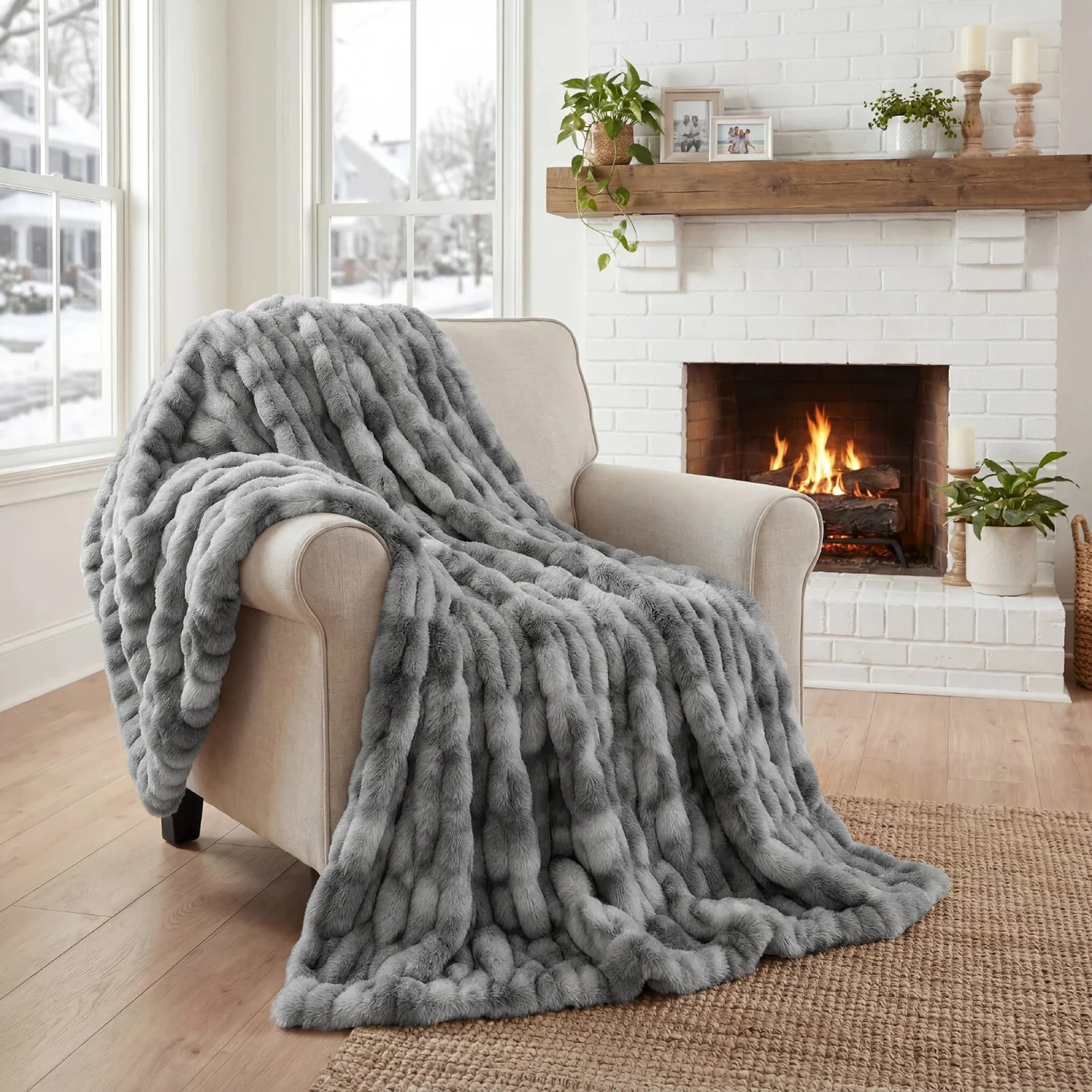A gray, ruched faux-fur throw blanket draped over a beige armchair next to a lit fireplace.