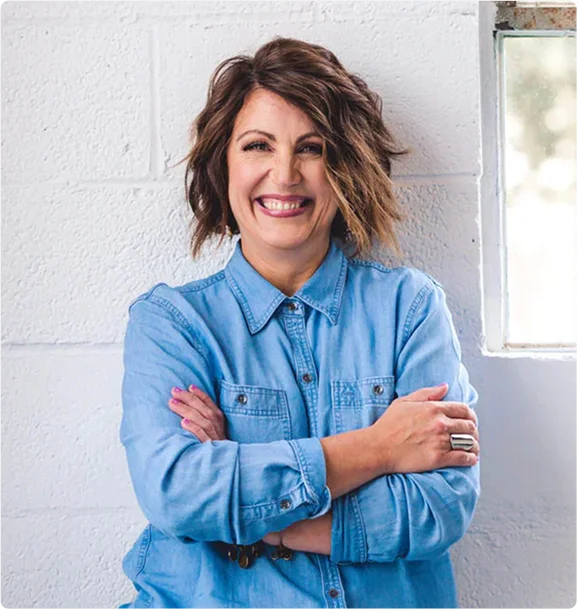 Smiling person in a blue denim shirt standing against a white brick wall.