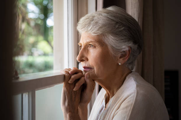 An elderly woman with short grey hair looks thoughtfully out of a window with her hands clasped.