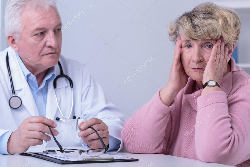 A distressed elderly woman holds her head in her hands while sitting with a male doctor.