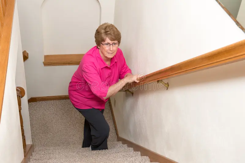 An older woman in a pink shirt struggles to walk up a carpeted staircase while holding the handrail.
