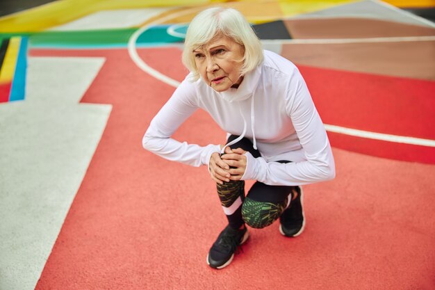 An elderly woman in athletic wear holds her knee with a pained expression on a colorful outdoor court.