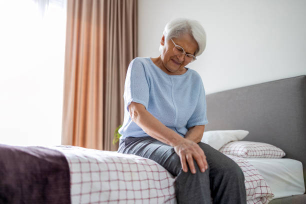 An elderly woman sits on the edge of a bed, holding her knee with a pained expression.