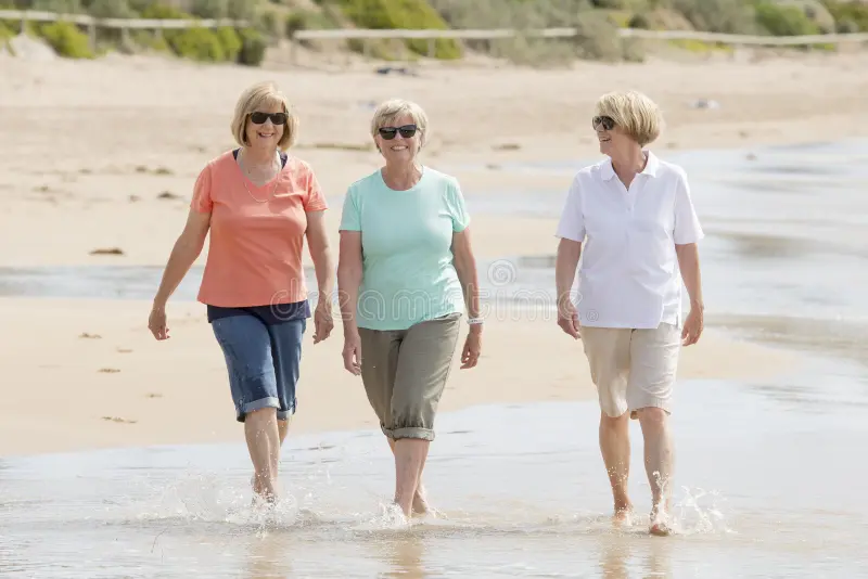 Three older women smiling while walking barefoot along the shoreline of a sandy beach.