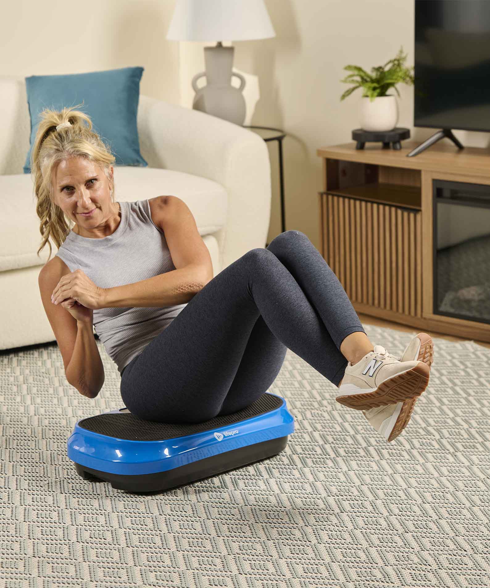 A woman in workout clothes does an abdominal exercise on a blue vibration plate in a living room.