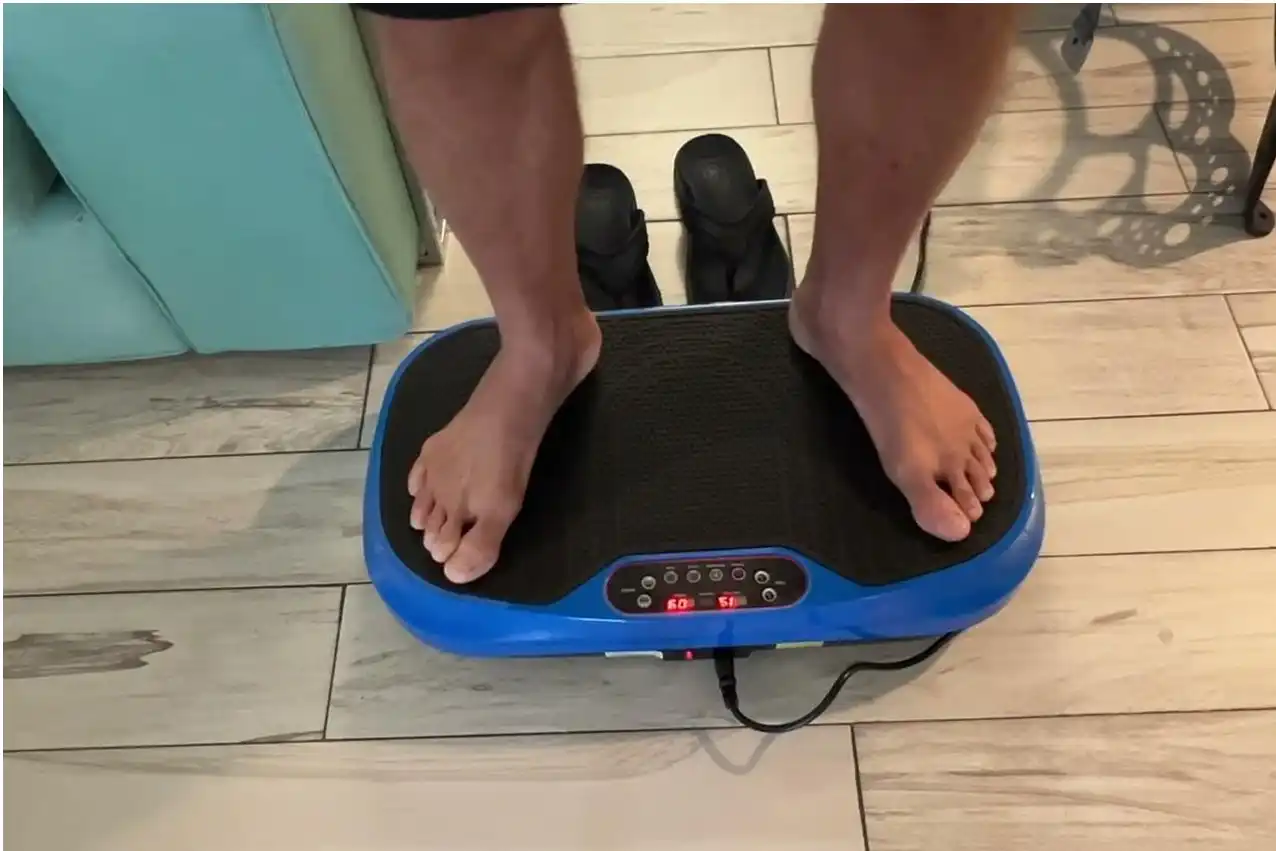 A person stands barefoot on a blue and black vibrating exercise platform on a tiled floor.