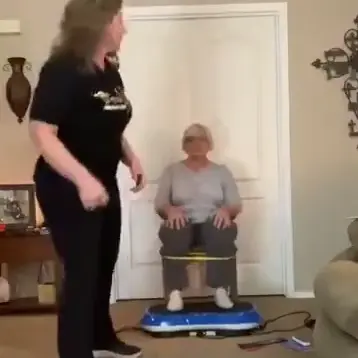 Two women in a living room, one sitting on a vibrating platform.