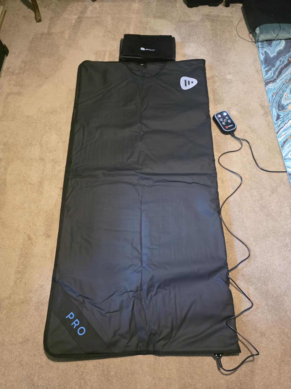 A black sauna blanket is laid out on a carpeted floor with its wired controller beside it.
