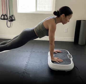 A woman in a plank position with her hands on a white vibration plate machine.