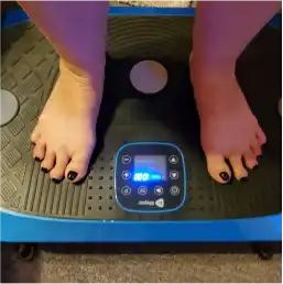 A person's feet with black painted toenails standing on a blue vibration plate exercise machine.