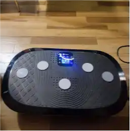 A black vibration plate exercise machine with a lit blue display sits on a light wood floor.
