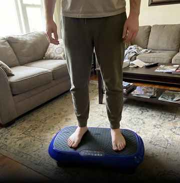 A person stands barefoot on a blue vibrating exercise platform in a living room.