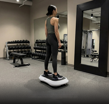 A woman in workout clothes stands on a vibration plate machine in a gym.