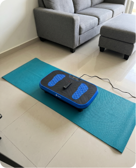 A blue and black vibration plate exercise machine sits on a teal mat in front of a grey sofa.