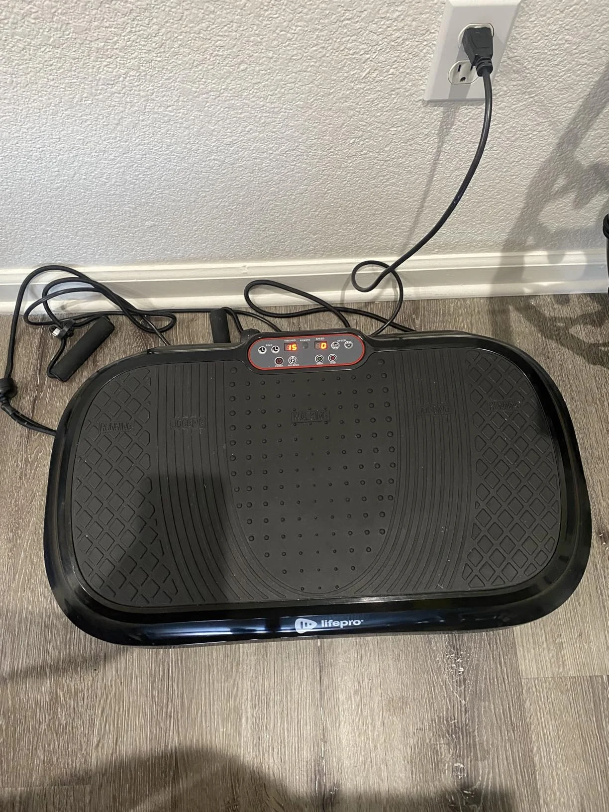 A vibrating exercise platform on a wooden floor, plugged into a wall outlet.