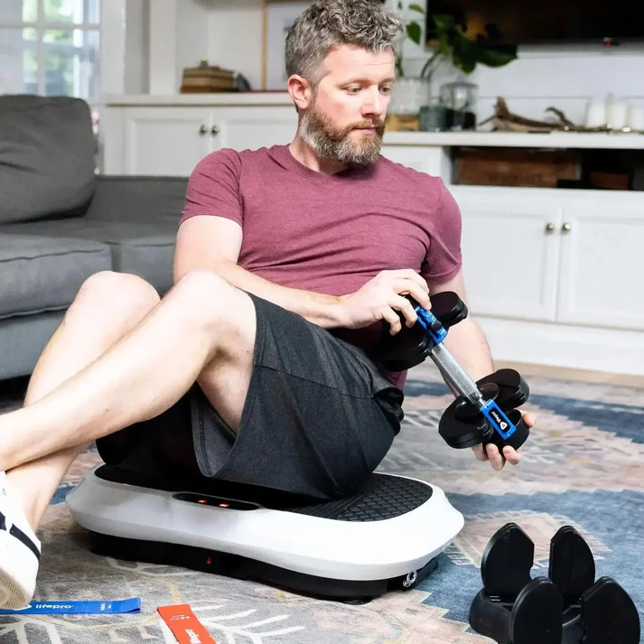 Person sitting on a vibration platform, adjusting a dumbbell in a living room.