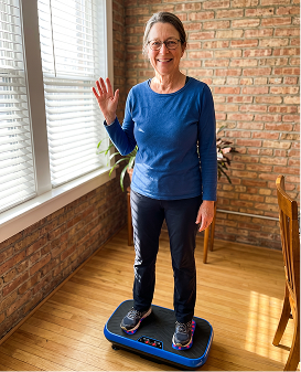 A woman in a blue shirt smiles and waves while standing on a vibration plate exercise machine indoors.