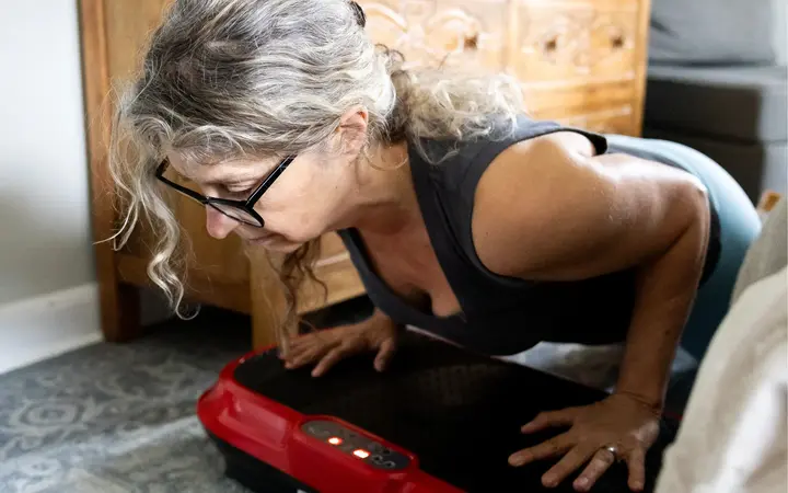 A woman with gray hair and glasses does a push-up on a red vibration plate exercise machine.