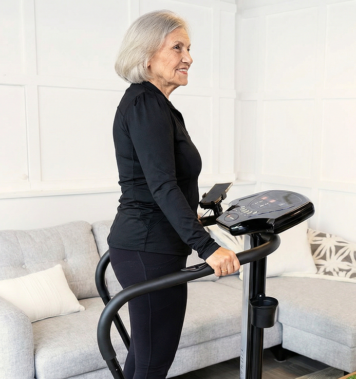 A woman with gray hair in black workout attire smiles while standing on an exercise machine at home.