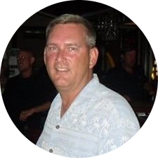 A circular photo of a smiling, middle-aged man with short hair wearing a patterned button-down shirt.