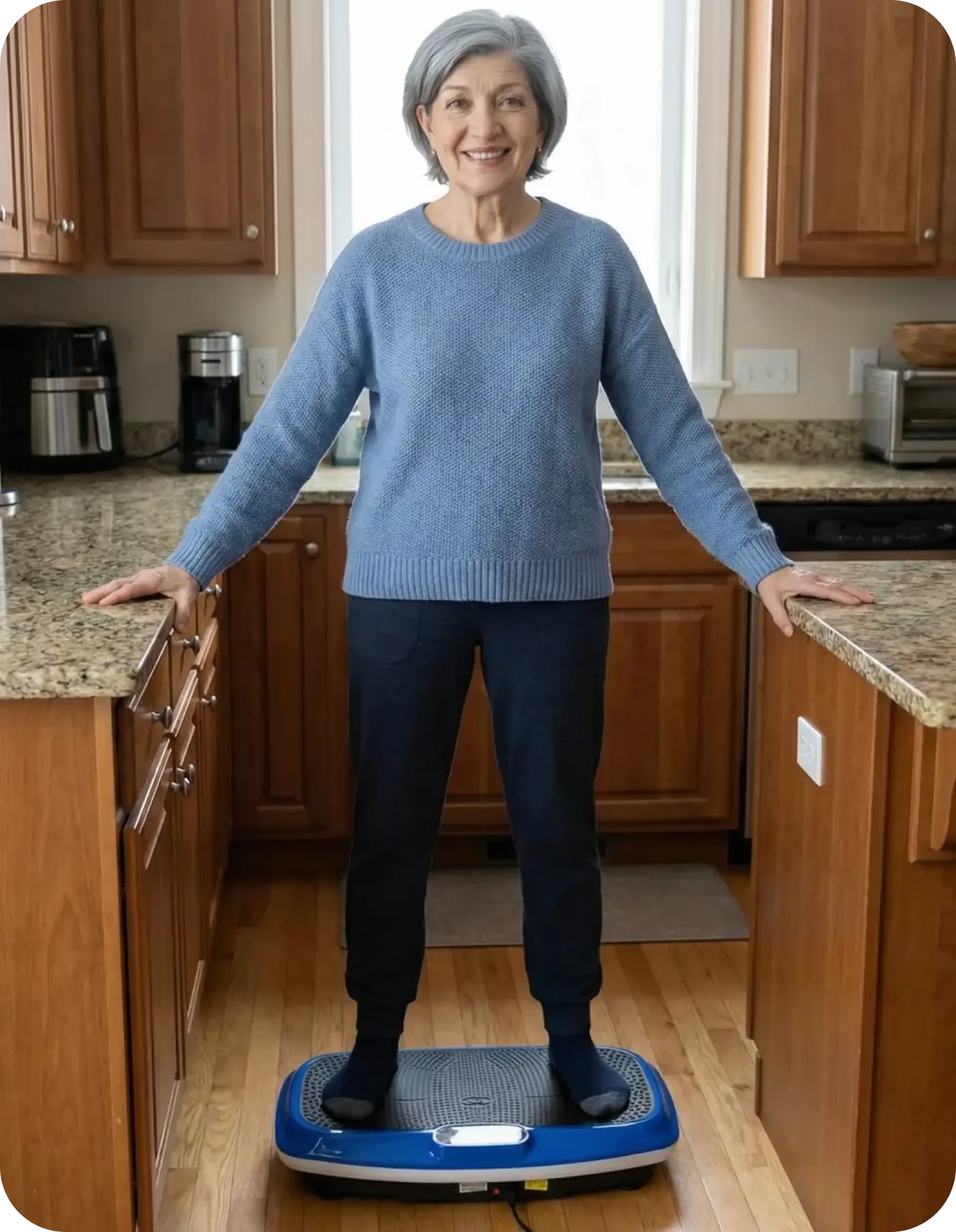 A woman with grey hair stands on a blue vibration plate in a kitchen, smiling at the camera.