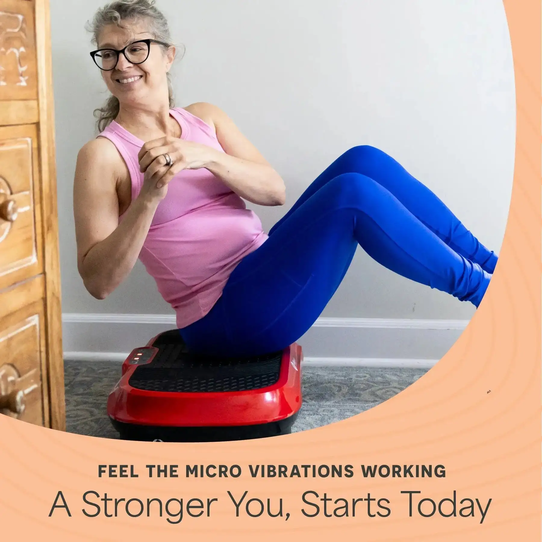 Person sitting on a vibration plate, wearing pink top and blue leggings, smiling.