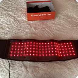A red light therapy belt with illuminated lights, lying on a white textured surface with its box behind it.