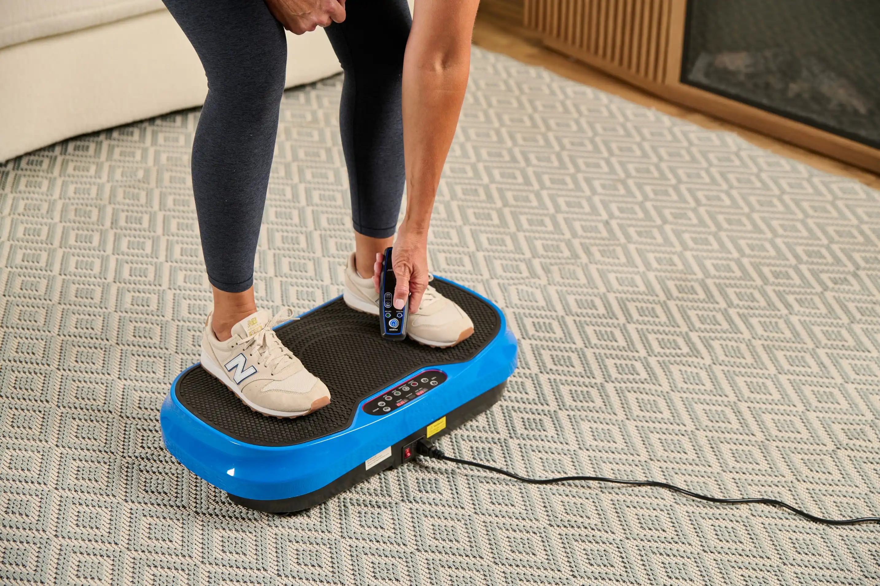 A person stands on a blue vibration plate exercise machine, holding the remote control.