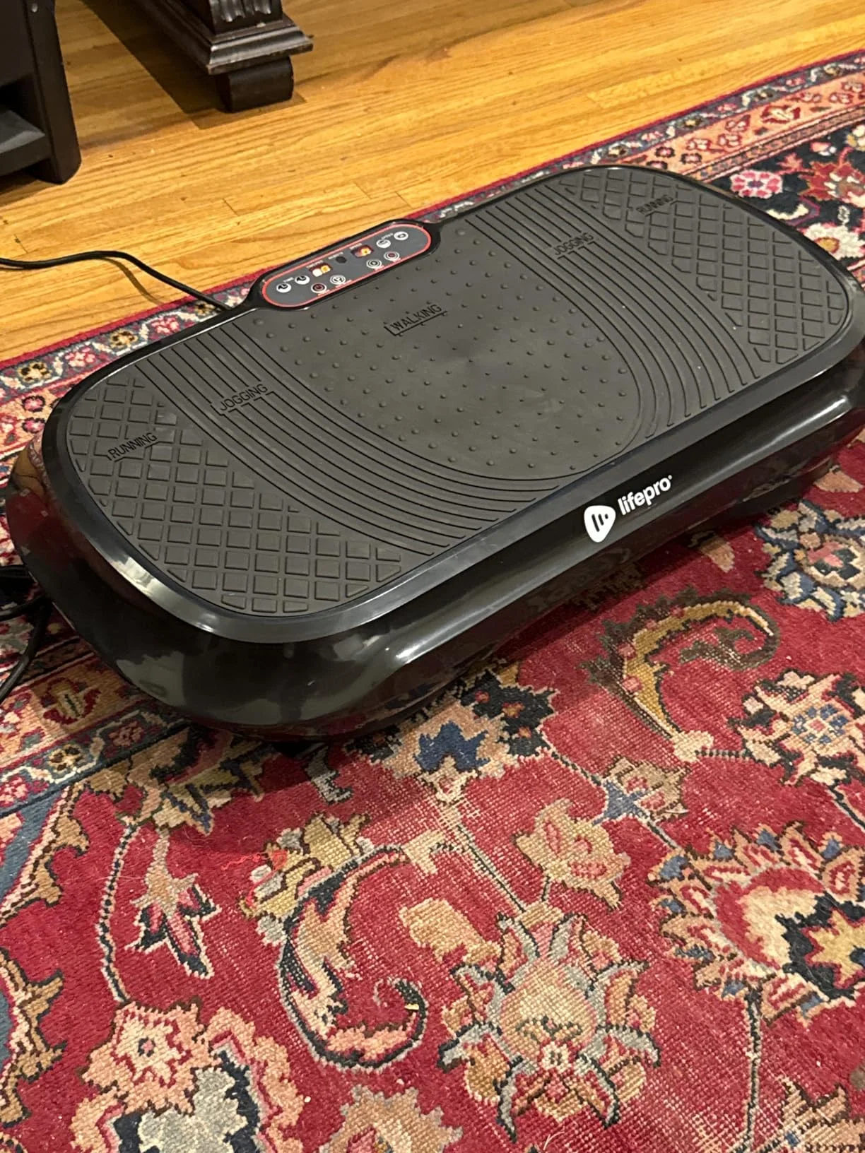 Black vibrating exercise platform on a patterned rug.