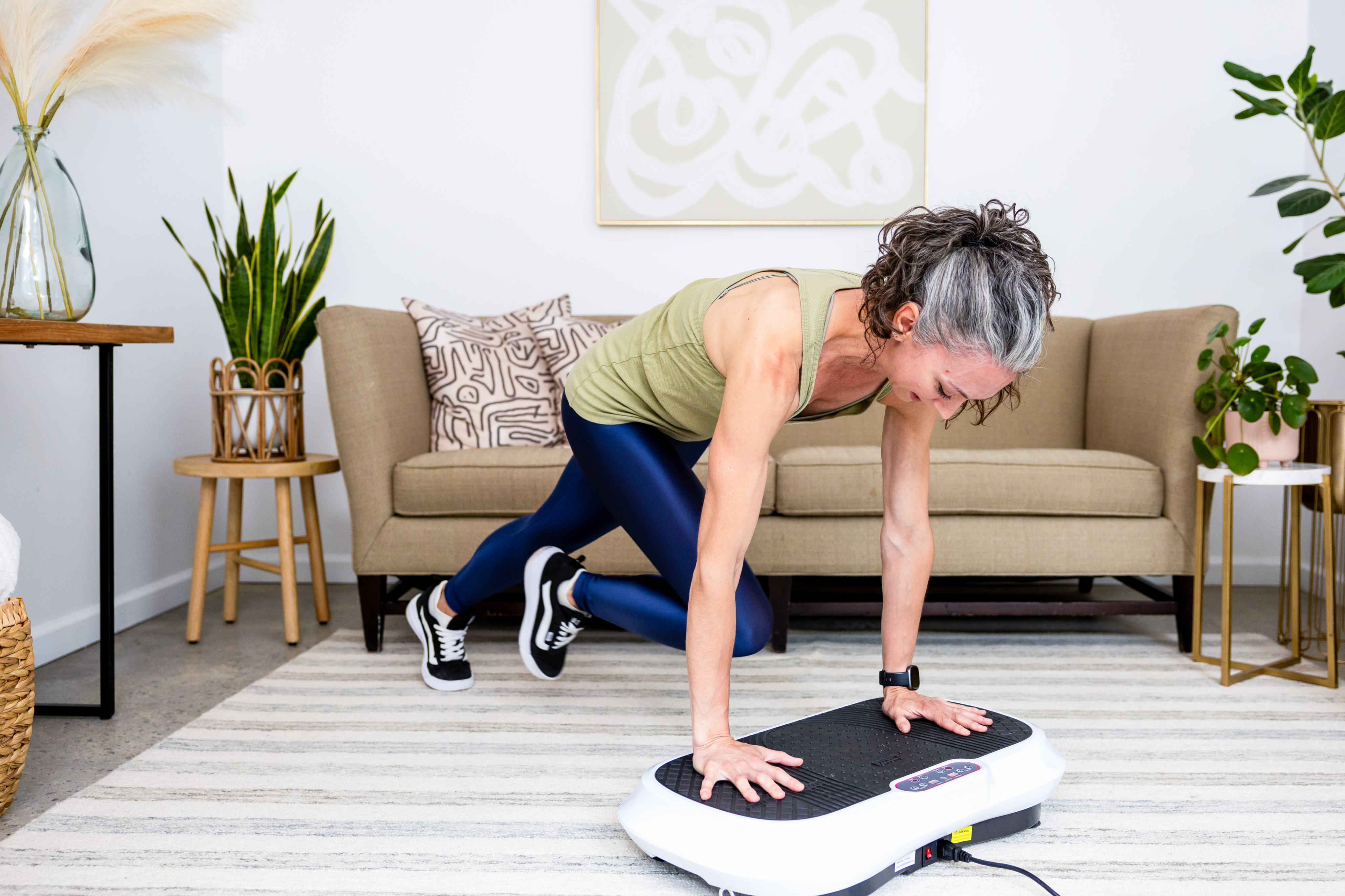 Person exercising on a vibration plate in a living room setting.