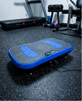 A blue and black vibration plate on a speckled gym floor, with a treadmill and weights behind it.