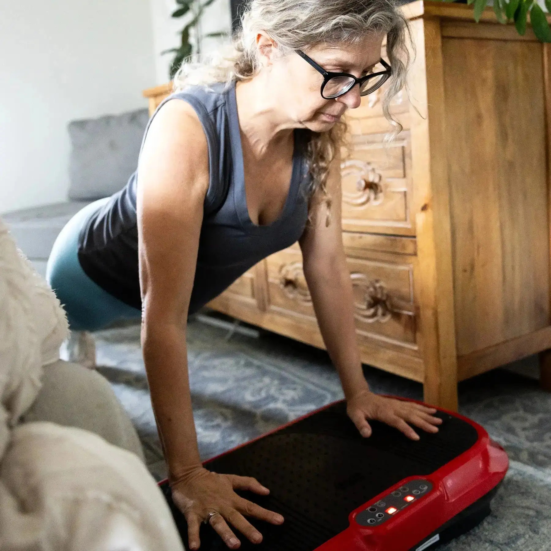 Person doing a plank on a red vibration plate indoors, near a wooden cabinet.