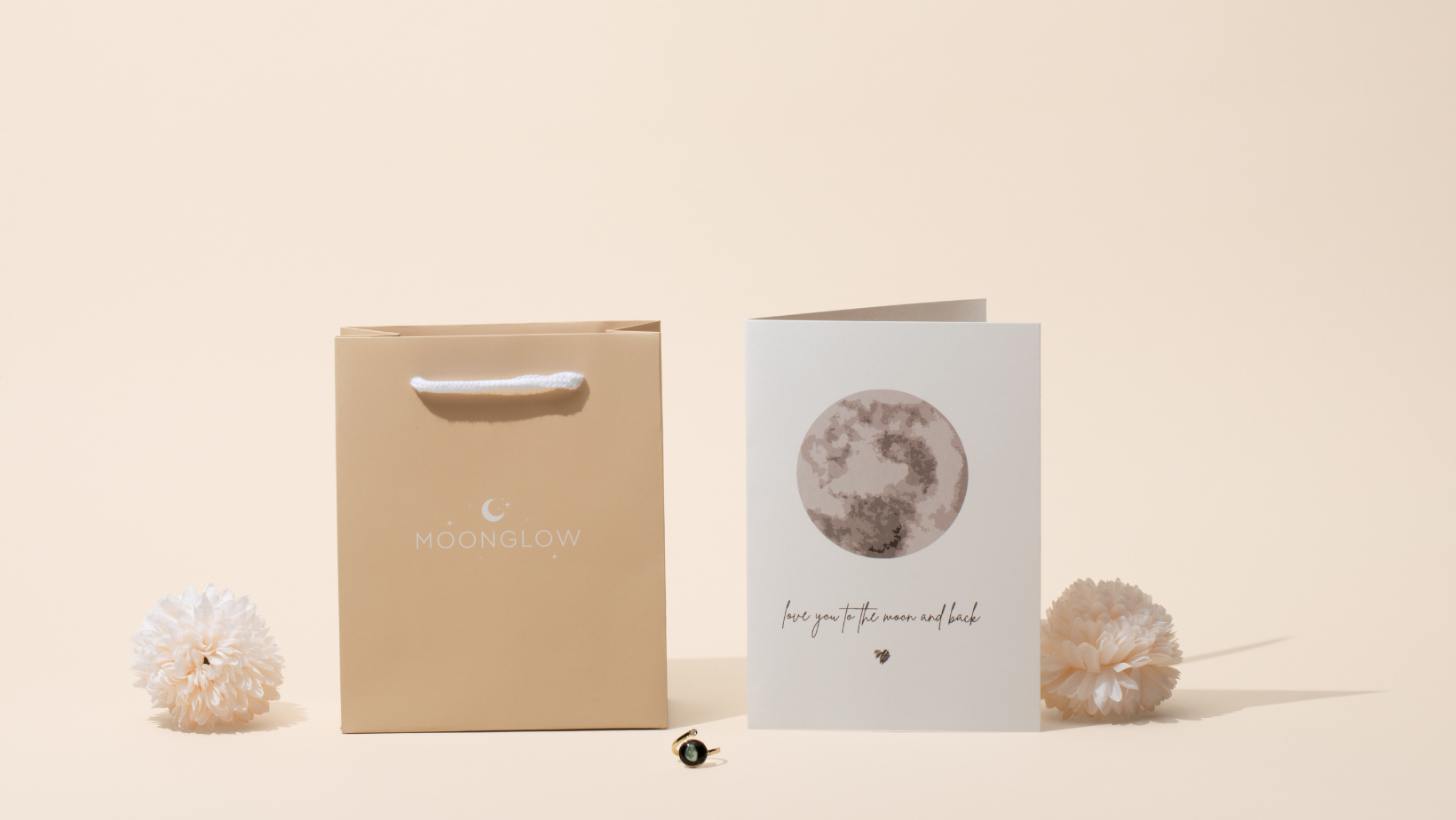 Gift bag, greeting card with moon design, and white flowers on beige background.