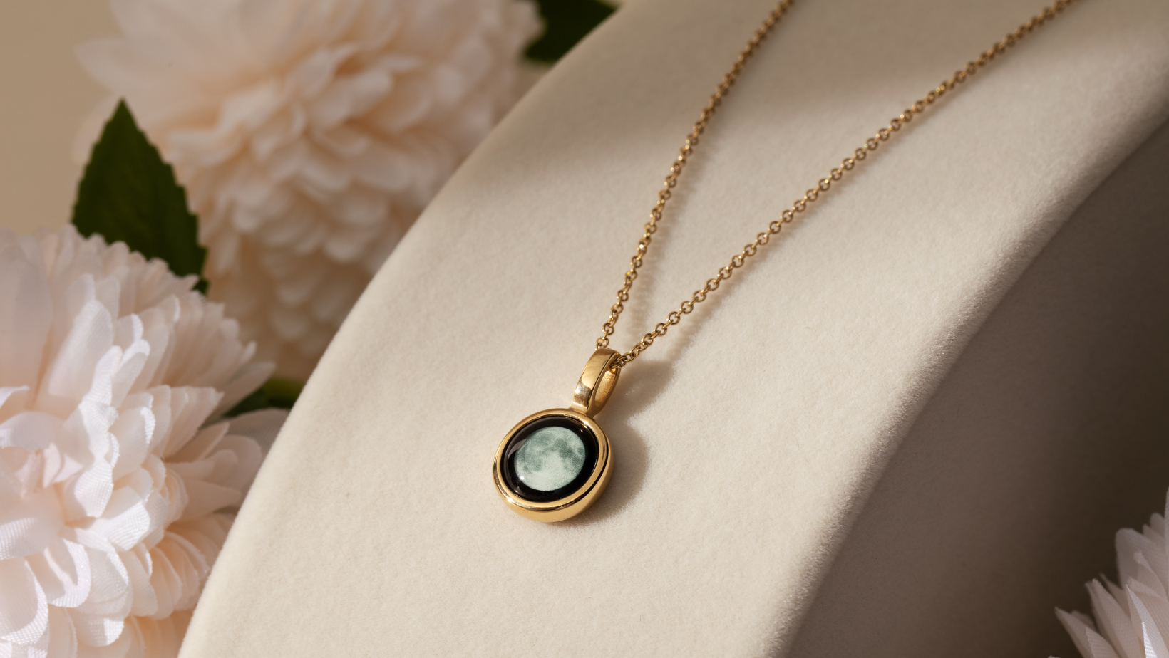 Gold necklace with moon pendant on a soft beige background, surrounded by white flowers.