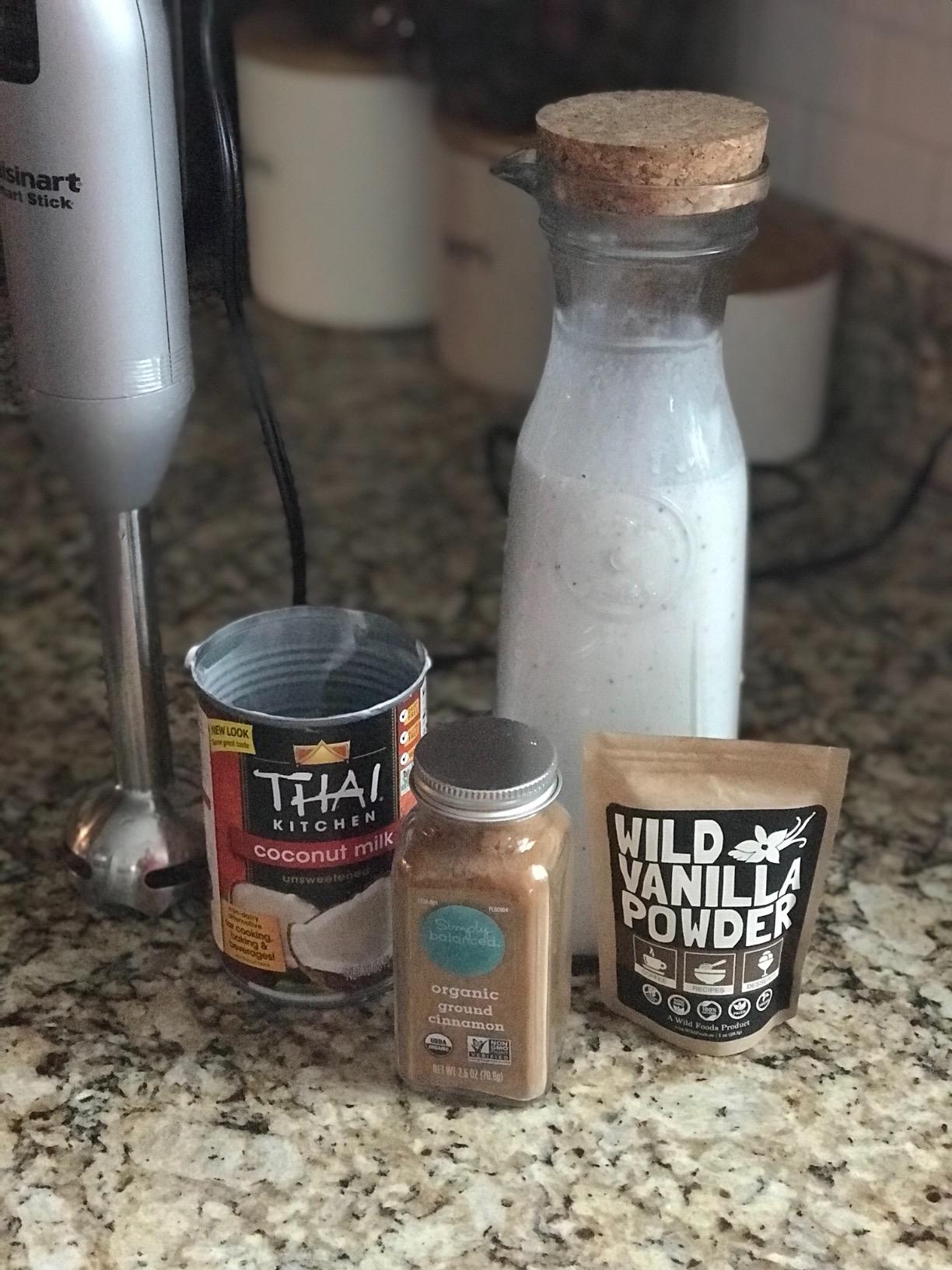 An immersion blender with coconut milk, cinnamon, vanilla powder, and a bottle of a white beverage.