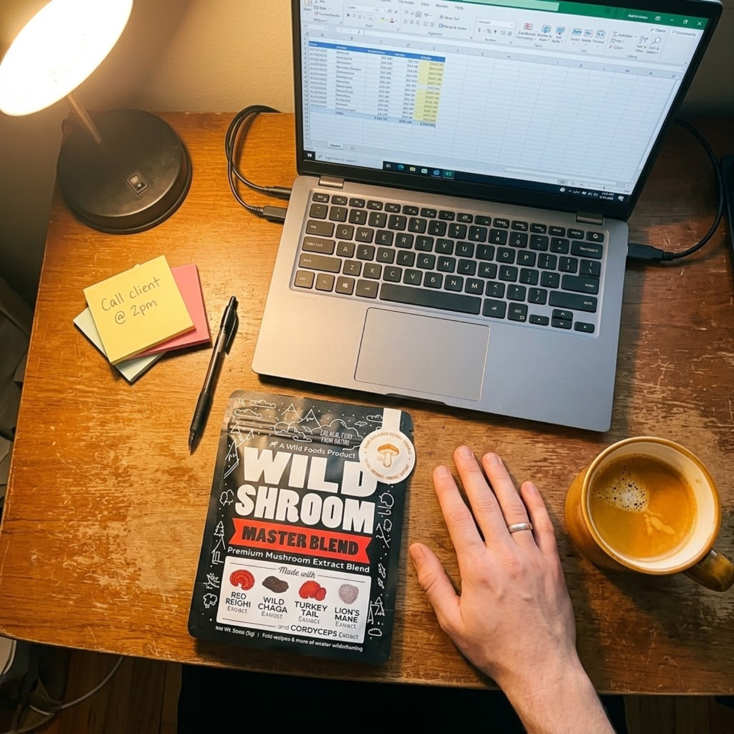 A top-down view of a desk with a laptop, a pouch of mushroom extract, a coffee mug, and a hand.