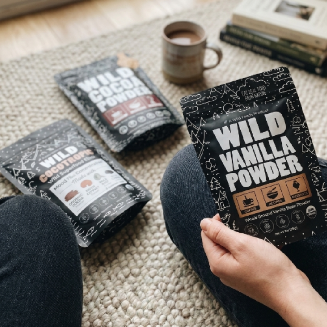 A person sits cross-legged on a rug, holding a pouch of Wild Vanilla Powder with other products nearby.