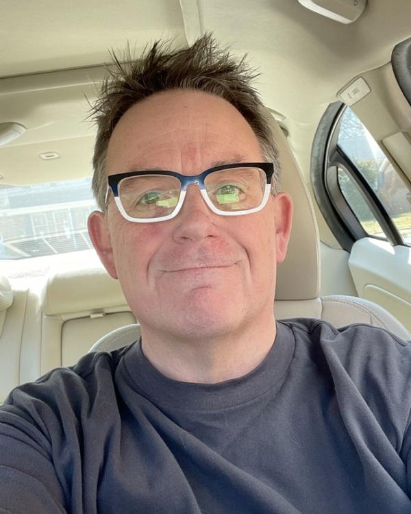 Person with glasses sitting in a car, smiling at the camera.