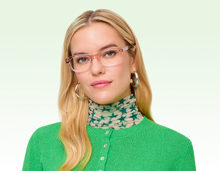 A woman with long blonde hair wears pink glasses, a green cardigan, and large silver hoop earrings.
