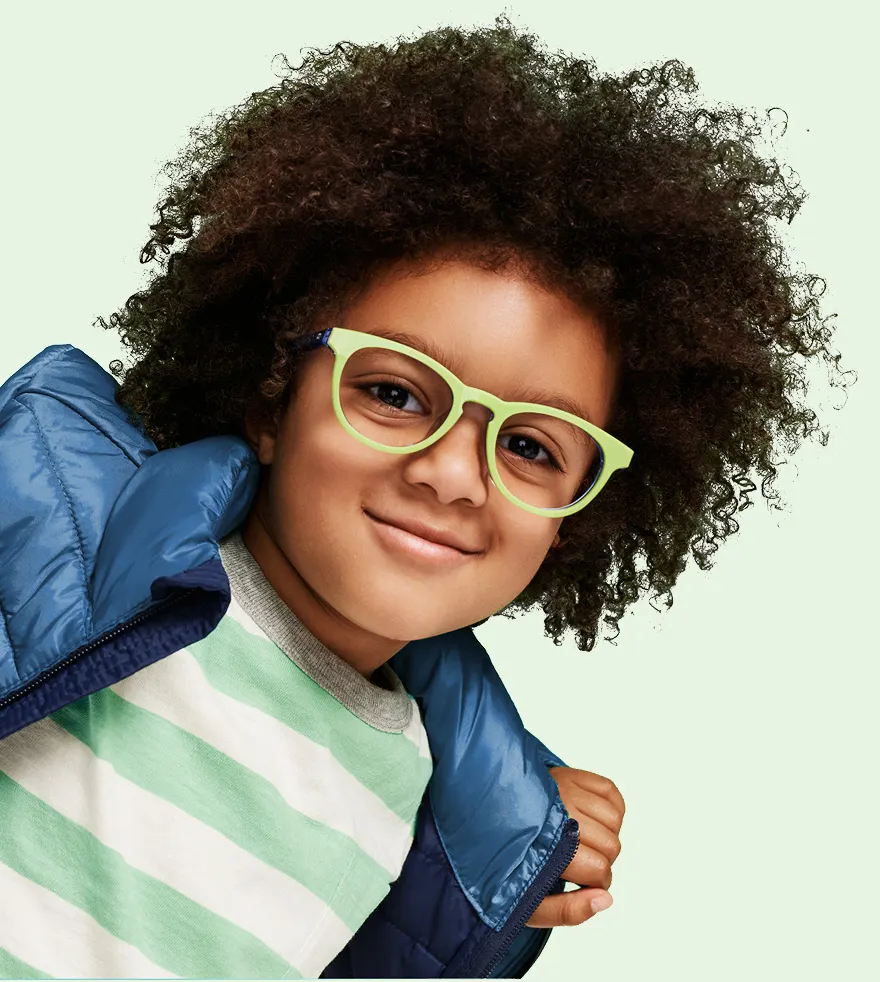 A smiling child with curly hair wears bright green glasses and a blue puffer jacket.
